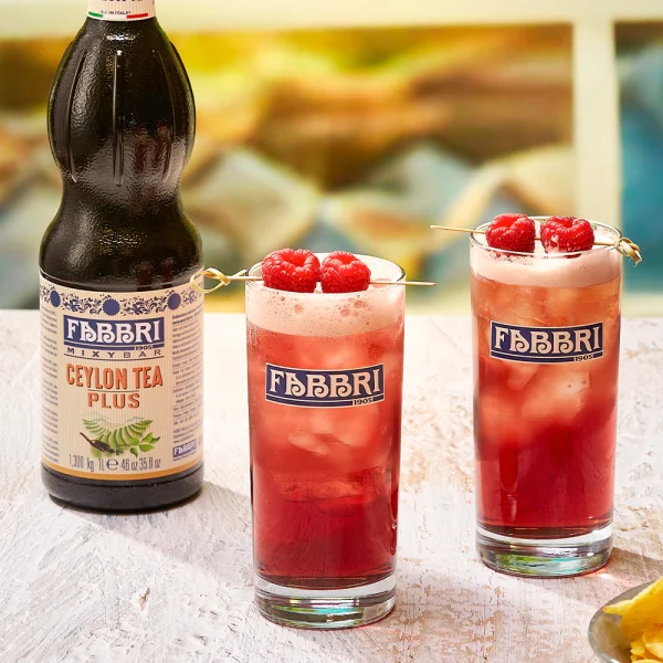 Uma garrafa de Xarope Fabbri Chá Preto 1 Litro Mixybar Ceylon Tea Plus Concentrado Profissional está ao lado de dois copos altos de chá vermelho gelado com espuma e framboesas, todos exibindo o logotipo da Fabbri.